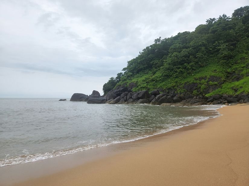 A beachside view with waves gently lapping at the shore, while half the frame is filled with lush, tree-covered mountains. The sky above is overcast, with clouds casting a soft, moody light