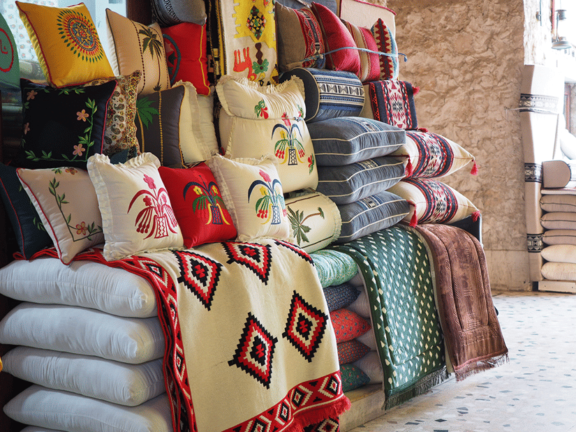 Colorful decorative cushions and embroidered patterned textiles are neatly stacked on display outside a shop store.