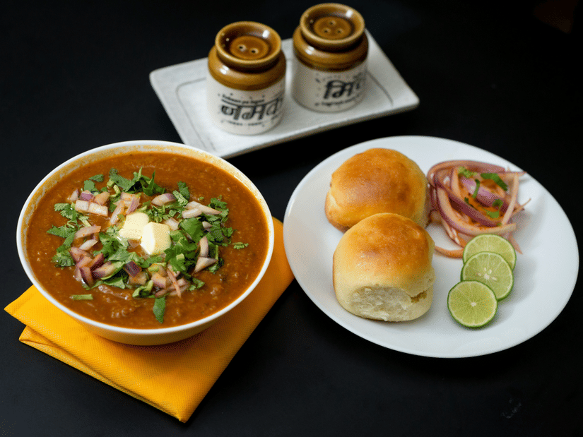 Bowl of pav bhaji topped with butter, chopped onions, and coriander, served with buttered pav buns, sliced onions, and lime wedges on a white plate.
