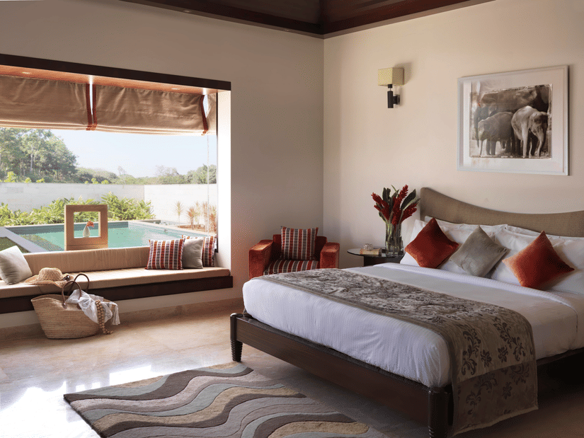 Interior of The Residence at The Serai Chikamagalur featuring a plush bed with a vase on the nightstand and next to is is a large window with seating space with a pool view
