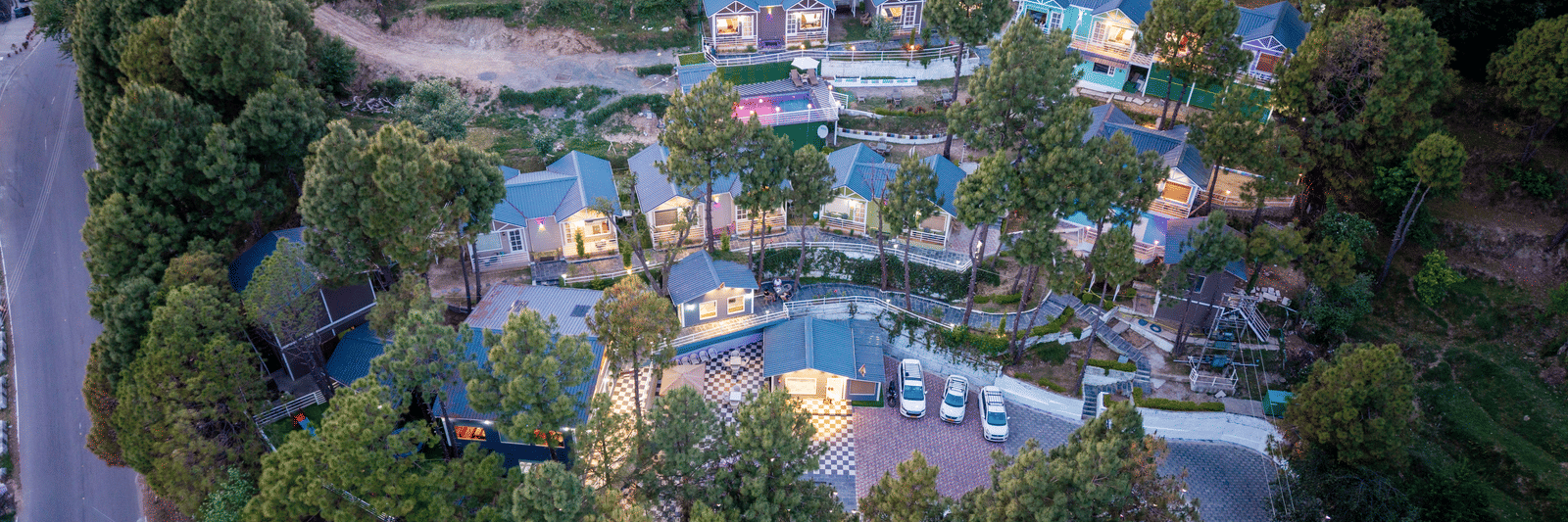 Aerial view of a scenic hillside resort featuring blue-roofed cottages, winding roads, and lush green trees, with nearby rural structures and natural surroundings - DLS Nature Trinket Resort, Dalhousie.