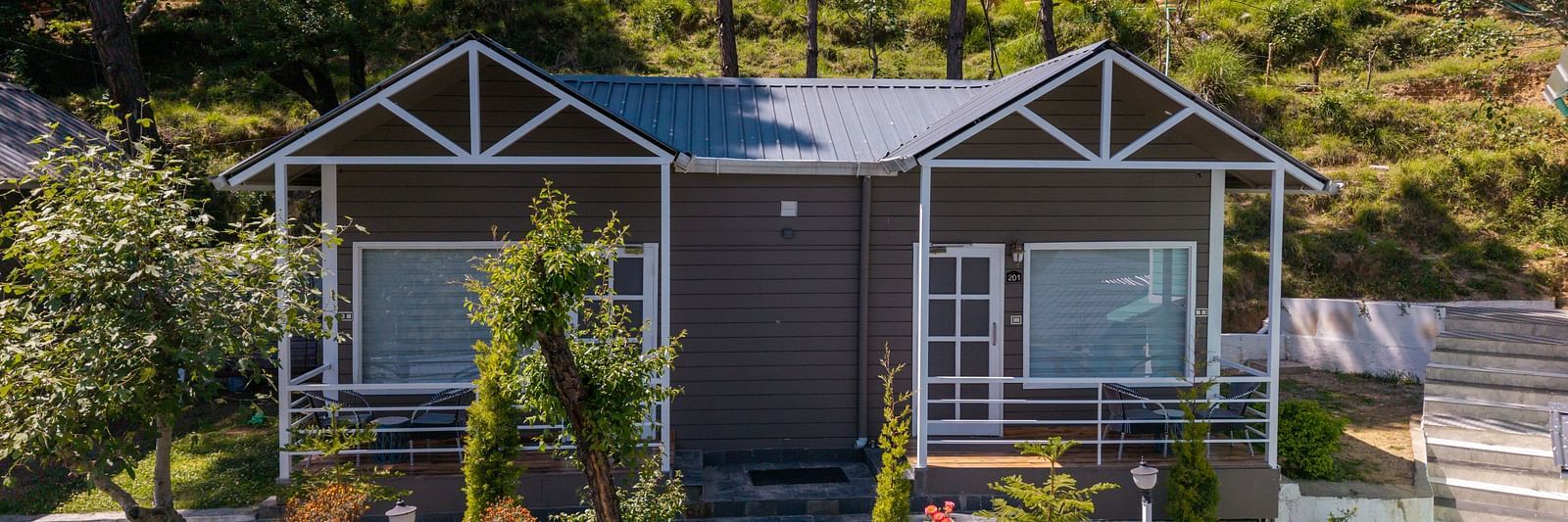 Charming hillside cottage with a twin-gabled roof, large front windows, and a landscaped garden surrounded by trees and greenery - DLS Nature Trinket Resort, Dalhousie.