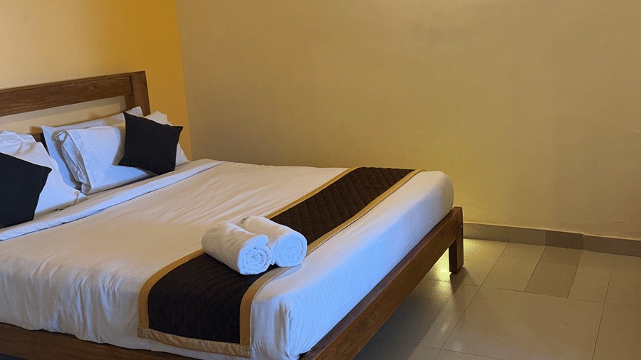 A bedroom with a wooden bed frame, white bedding, and a small side table covered with a patterned red tablecloth | Kollanoor Guest House, Coorg