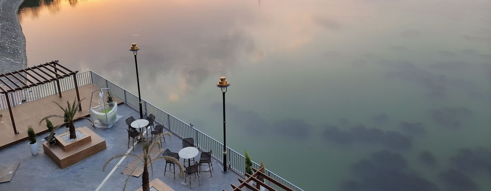 An outside pool area view of a lake surrounded by hills, with a seating area and a walkway along the shore, during the sunset  time.