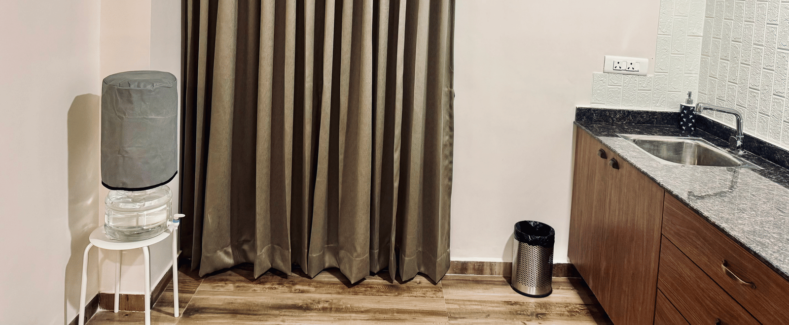 Kitchen of Coral 4BHK at Kove Smart Stays furnished with a separate water facility placed in front of closed curtains, and a countertop on the other side.