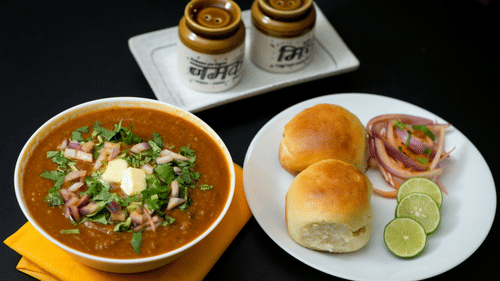 A plate of pav bhaji served with bread rolls, onion slices, lemon wedges, and two cups of tea on a table.