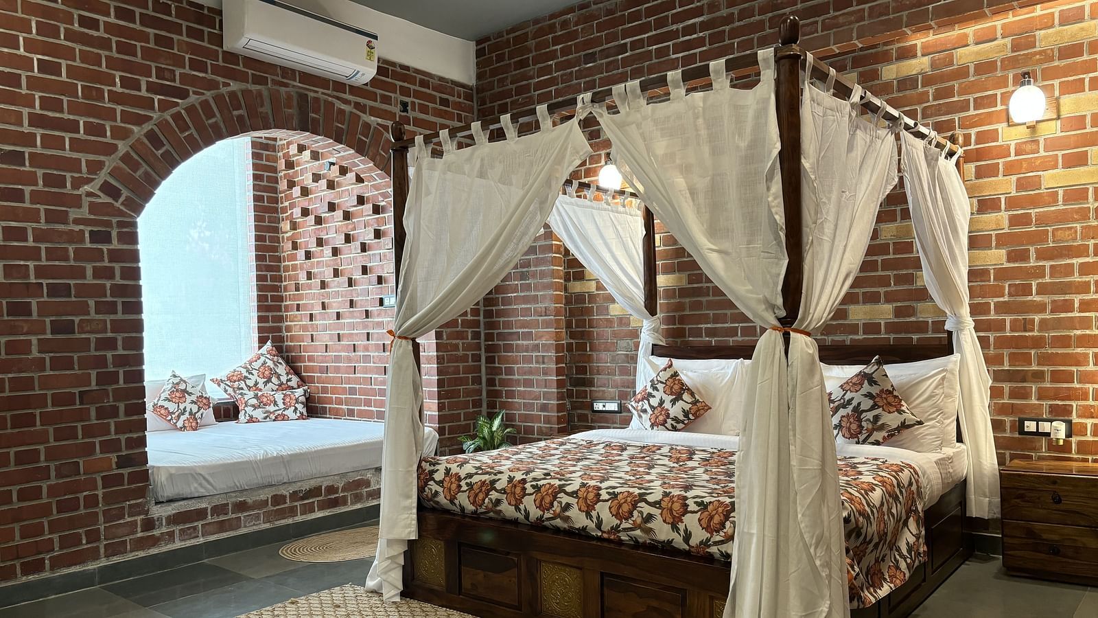 A bedroom at Tisya Farms, Gurgaon with a four-poster bed, white curtains, brick walls, and an arched window.