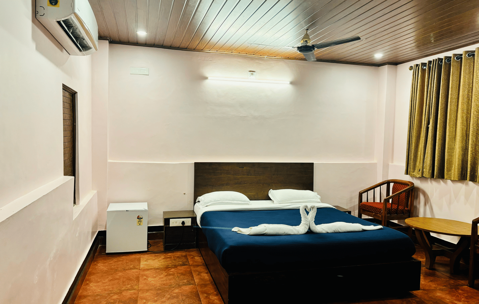 Bedroom with a queen bed, air-conditioning, a wooden table and chairs, a mini fridge and green curtains at Paradise Lagoon Resort, Udupi.