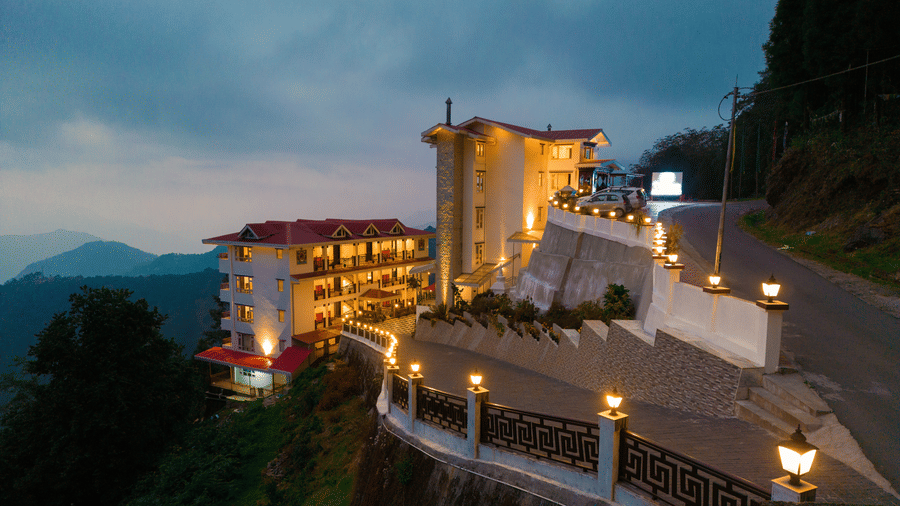 Scenic view of a hilltop hotel with glowing lights, red-roofed buildings, and mountain backdrops at dusk.
