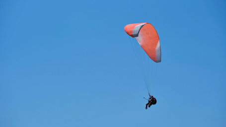 A person glides through the air using a large canopy wing against a clear blue sky.