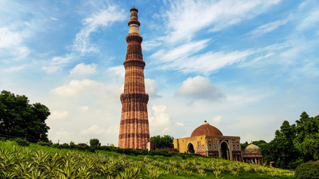 Qutb Minar rises above landscaped gardens in Delhi, showcasing Indo-Islamic architecture beneath dramatic clouds and bright daylight, one of the popular places to explore on a Delhi Day Tour