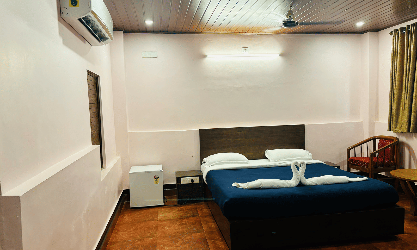 A wide angle view of a bedroom with a queen bed, a mini fridge,  a wooden table and chairs, and green curtains at Paradise Lagoon Resort, Udupi.