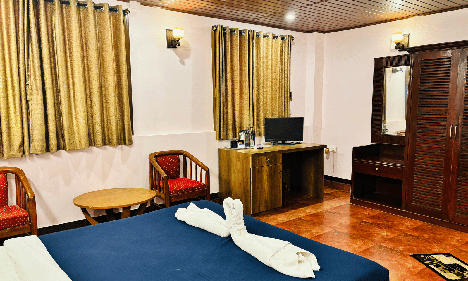 A partially visible bed with decorative towel, wooden table and red chairs, wooden cabinet and a television, a wardrobe and a dressing mirror at Paradise Lagoon Resort, Udupi.