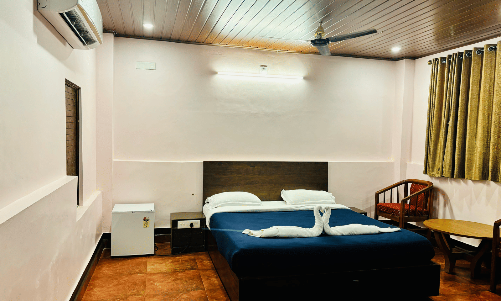 Bedroom with a queen bed, air-conditioning, a wooden table and chairs, a mini fridge and green curtains at Paradise Lagoon Resort, Udupi.
