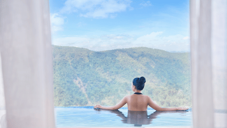 Woman relaxes in an infinity pool at Polo Resort Cherrapunji, leaning on the edge while gazing at forested rolling hills under a partly cloudy blue sky.