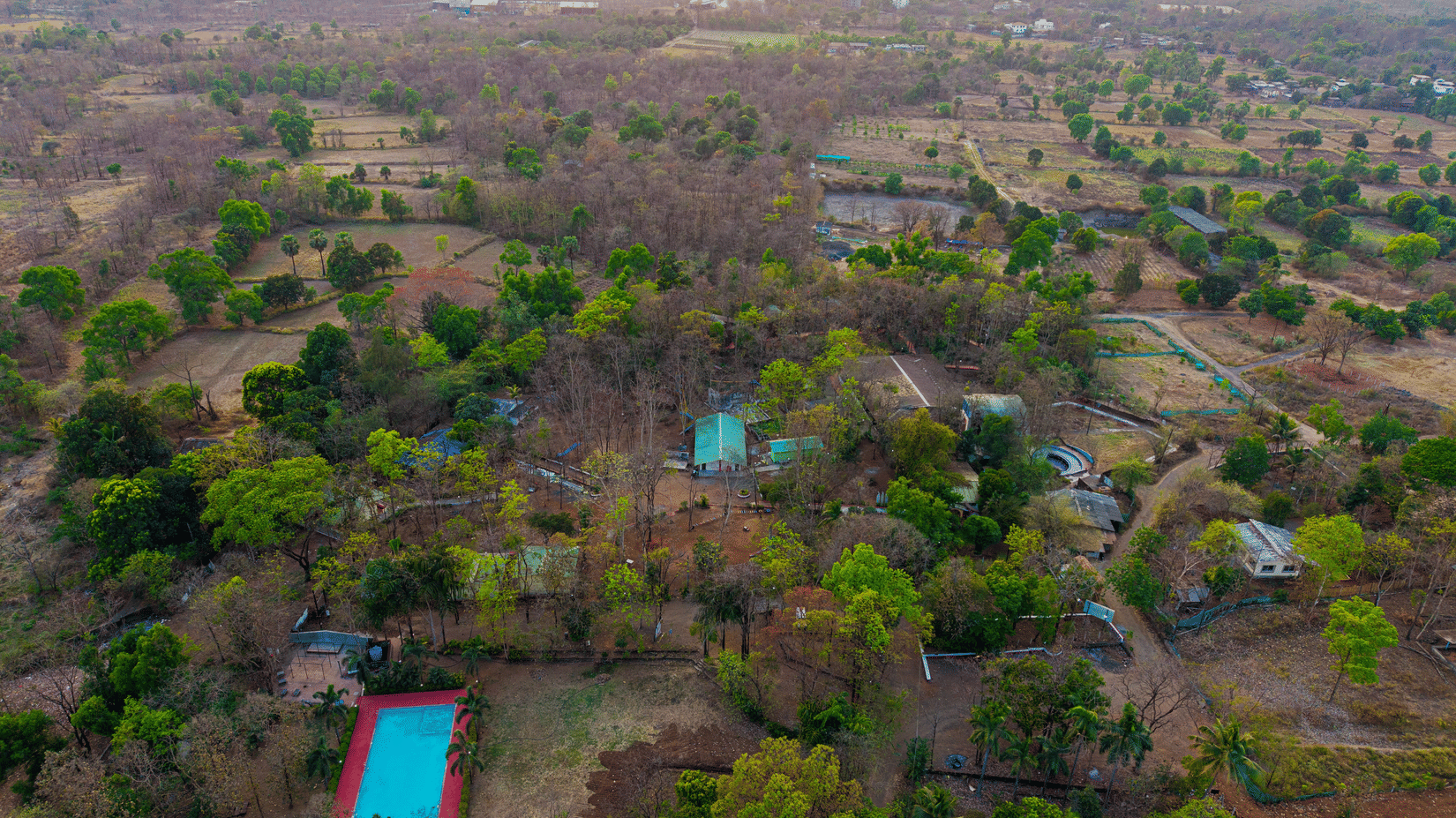 Aerial drone view of Nature Trails Sajan resort surrounded by dense greenery, featuring a blue swimming pool, red-roofed buildings, and vast landscape.