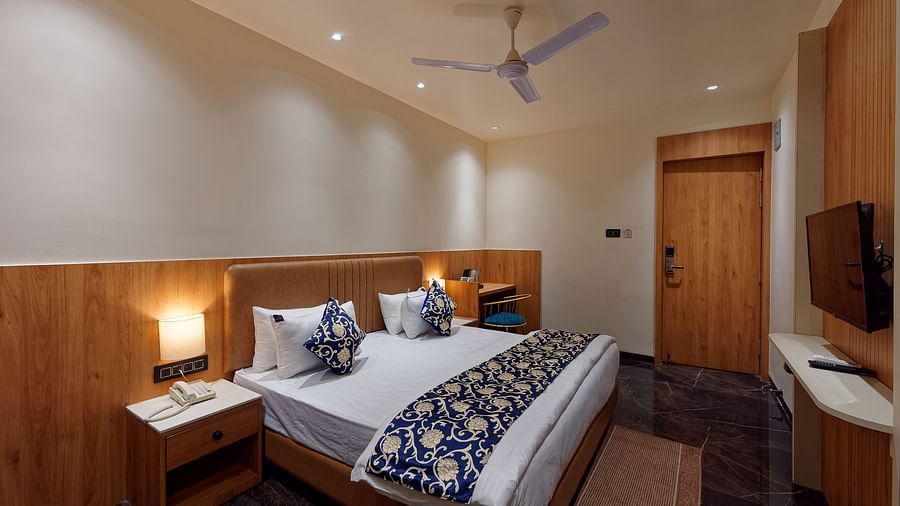 Image of a studio room with a bed with a table on the side of the bed and a television in front Image of a bed with table and chair on the side of the table and a fan on te ceiling at Ankit Hotel, Jabalpur