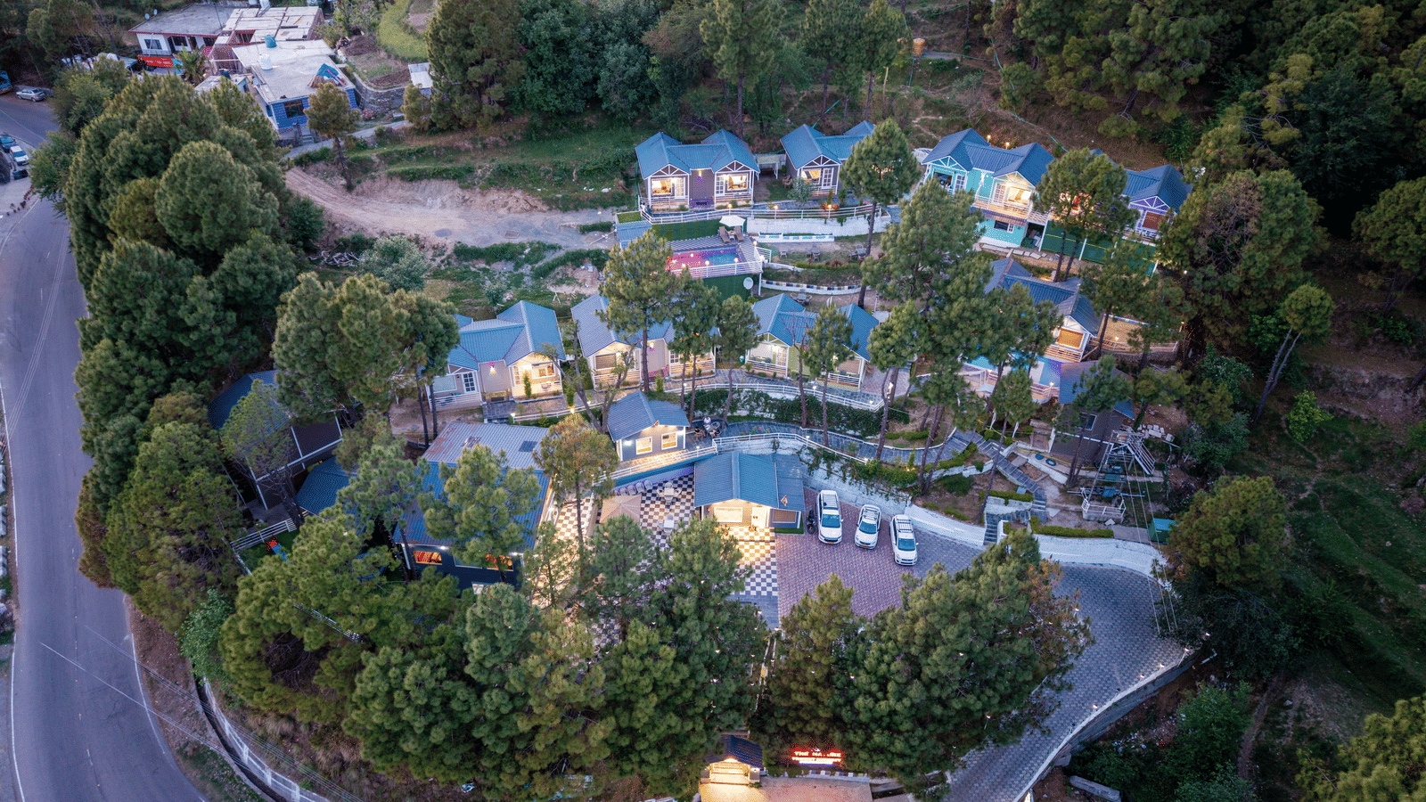 Aerial view of a scenic hillside resort featuring blue-roofed cottages, winding roads, and lush green trees, with nearby rural structures and natural surroundings - DLS Nature Trinket Resort, Dalhousie.
