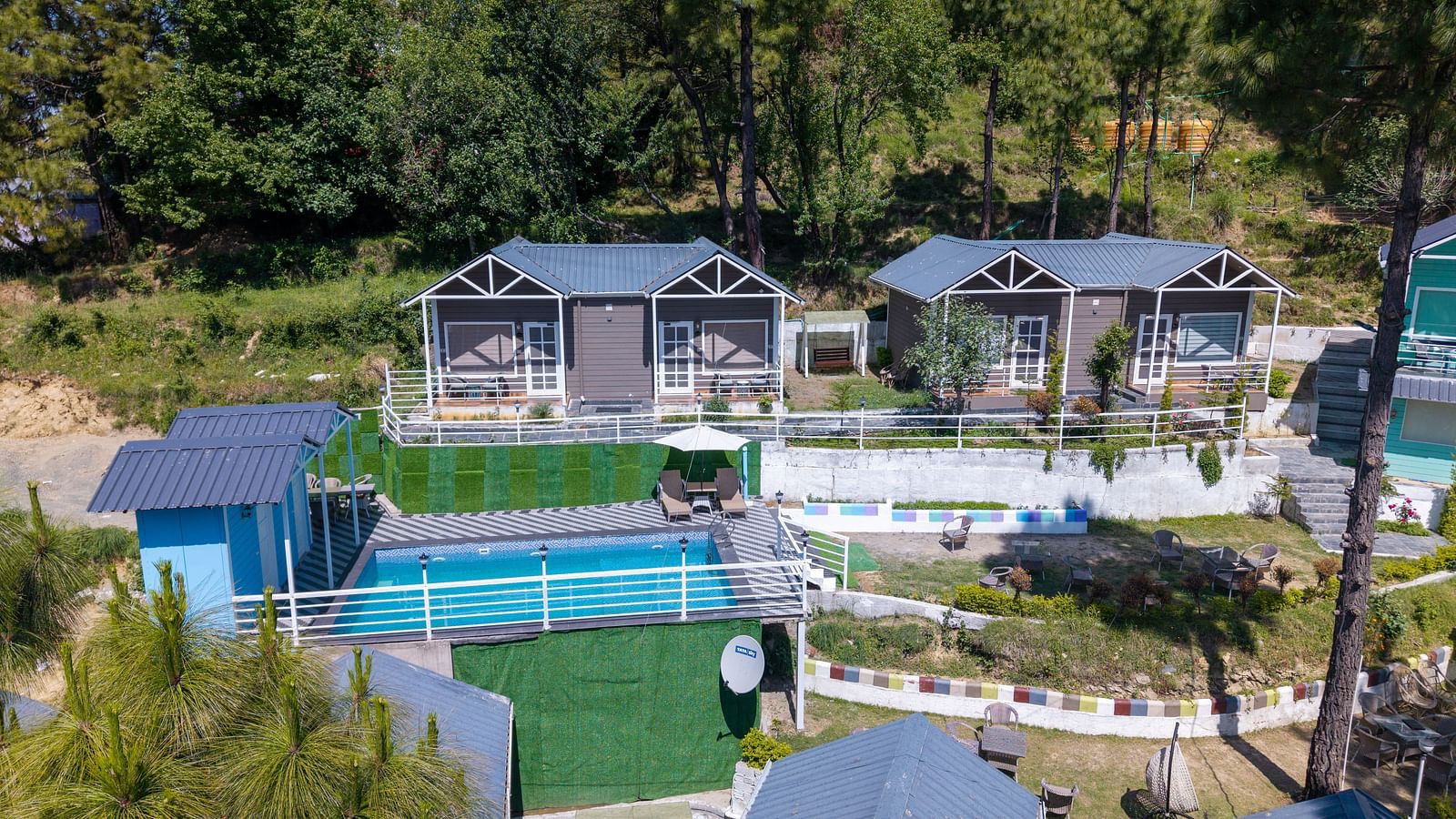 High-angle shot of two modern cabins with large windows, situated on a terraced hillside overlooking a blue swimming pool. Lush green trees surround the property - DLS Nature Trinket Resort, Dalhousie.