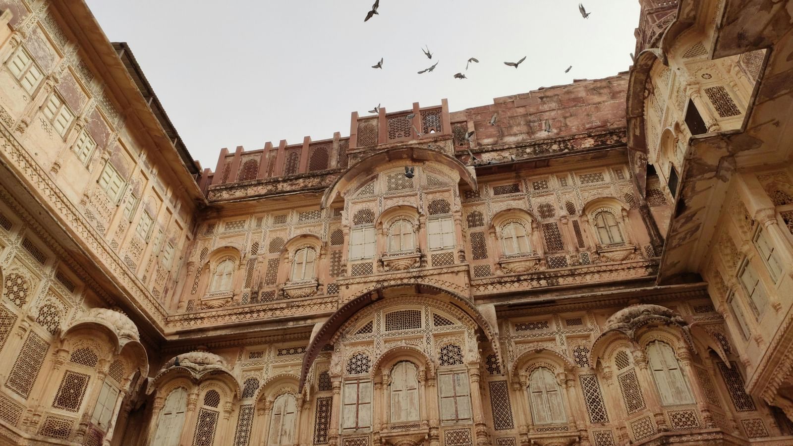  A low-angle view of the intricately carved sandstone facade of an historic Indian palace or fort, with multiple ornate windows and a flock of birds flying overhead.