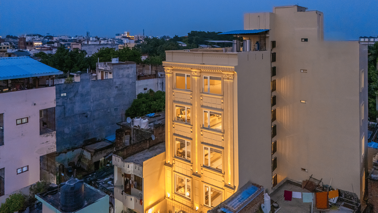 Aerial view of Perfectstayz Premium Lavender, Varanasi at dusk, a cream 5-storey building with lit windows and pillared facade among neighbouring rooftops and trees.