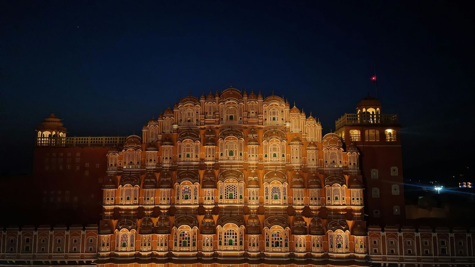 The Hawa Mahal in Jaipur is illuminated at night, showcasing its intricate facade and numerous small windows against a dark blue sky.