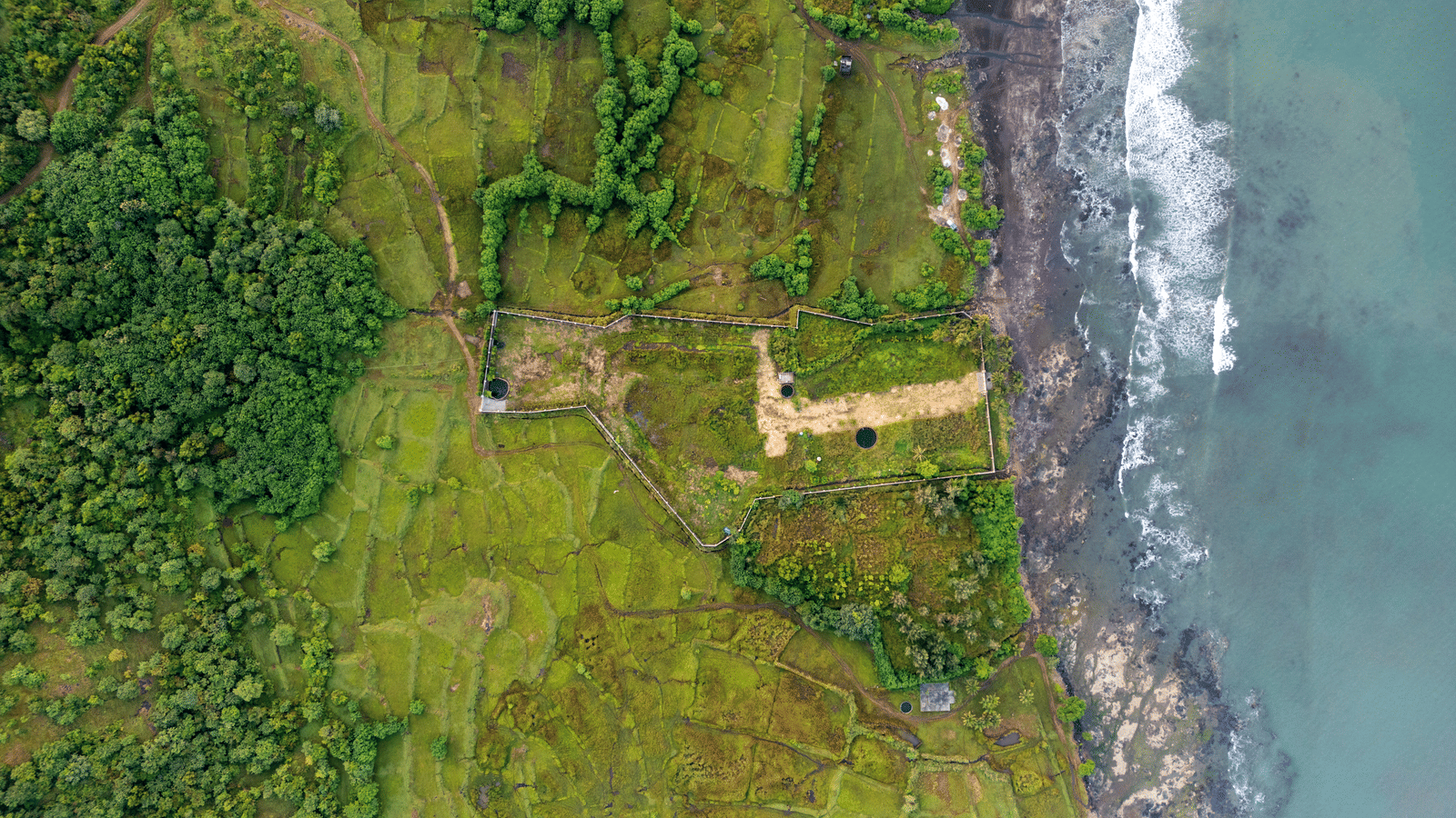 Top-down aerial shot of coastline, showing waves hitting rocky shore, sections of grassy land, and a rectangular ruin near the edge. Dense green areas cover much of the scene beside the water.