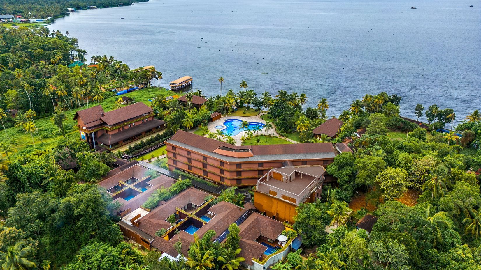 An aerial view of a hotel nestled among lush greenery, with a breathtaking expanse of the vast ocean and clear sky in the background - Uday Backwater Resort
