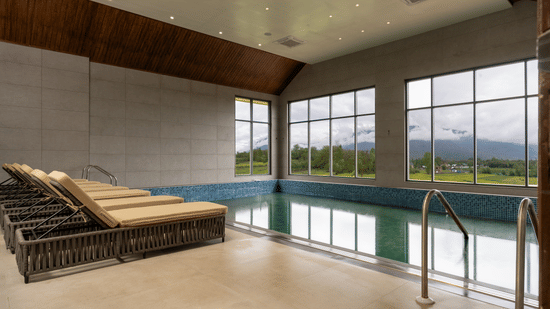 Indoor swimming pool at Mountnoor Sarovar Premiere, Pahalgam overlooking tranquil mountains through large glass windows.