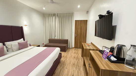 Wide-angle view of Executive Cottage at VITS Daman Devka Beach with king bed, purple headboard, sofa, TV unit, and wooden flooring in open layout.
