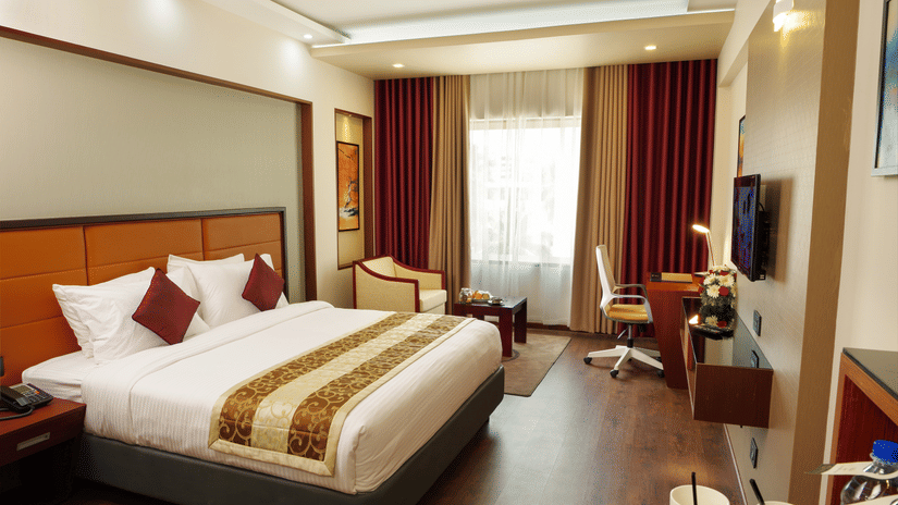 The Standard Room at Dimora, Thiruvananthapuram with a king size bed, a work desk, a flat screen television, a glass window with curtains, and wooden flooring.