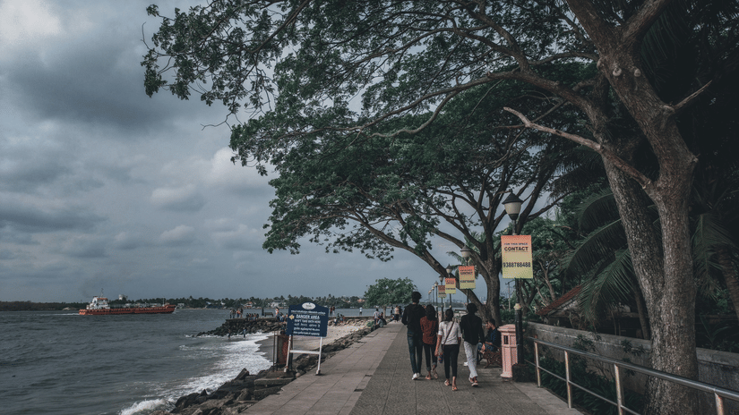 A tree-lined waterfront promenade curves beside the sea under overcast skies, with pedestrians strolling and distant ships visible offshore
