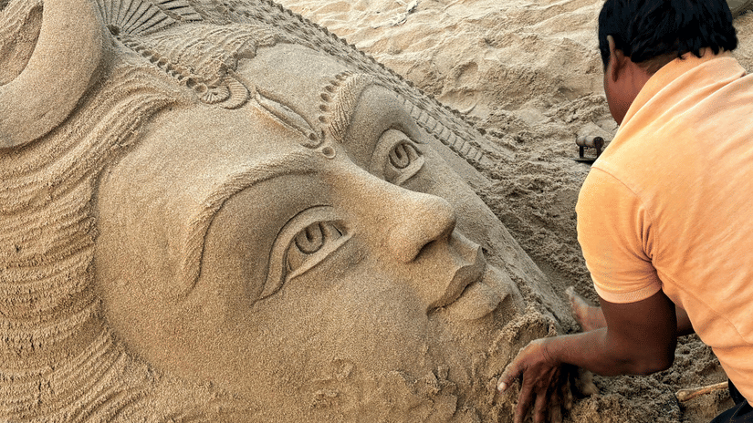 A person uses their hands to shape a large face into the sand on a beach. The sculpture features detailed eyes and a crown.
