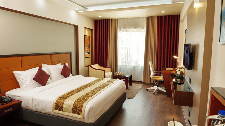 The Standard Room at Dimora, Thiruvananthapuram with a king size bed, a work desk, a flat screen television, a glass window with curtains, and wooden flooring.