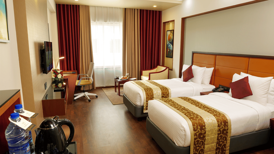 The Standard Room with twin beds at Dimora, Thiruvananthapuram, with twin beds, a flat screen television, glass window with curtains, and wodden flooring.