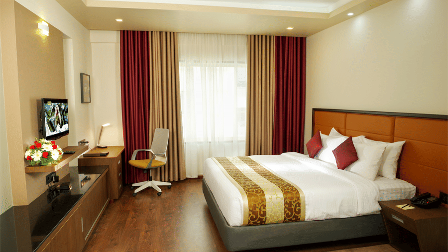 The Suite at Dimora, Thiruvananthapuram with a king size bed, a glass window with long curtains, ceiling lights, wooden flooring, and work desk.