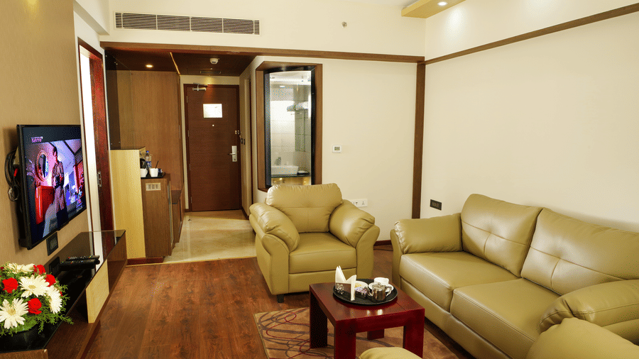 A Suite living area t Dimora Thiuvananthapuram with a multi-seating sofa, a rug, a coffee table, a flat screen television, wooden flooring, and an opening to another room.