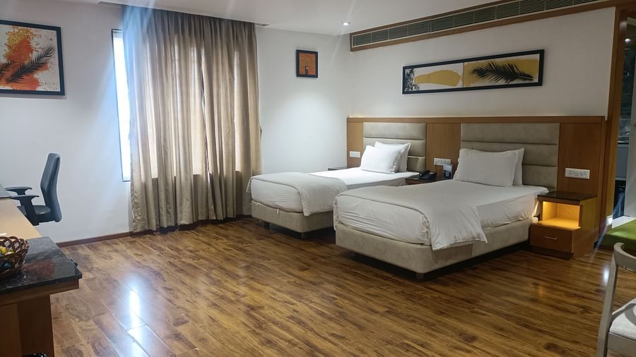 A wide-angle view of a Deluxe Twin room at Kyriad Hotel, Gulbarga, featuring two neatly made beds, dark wood flooring, and a framed abstract artwork on the wall.