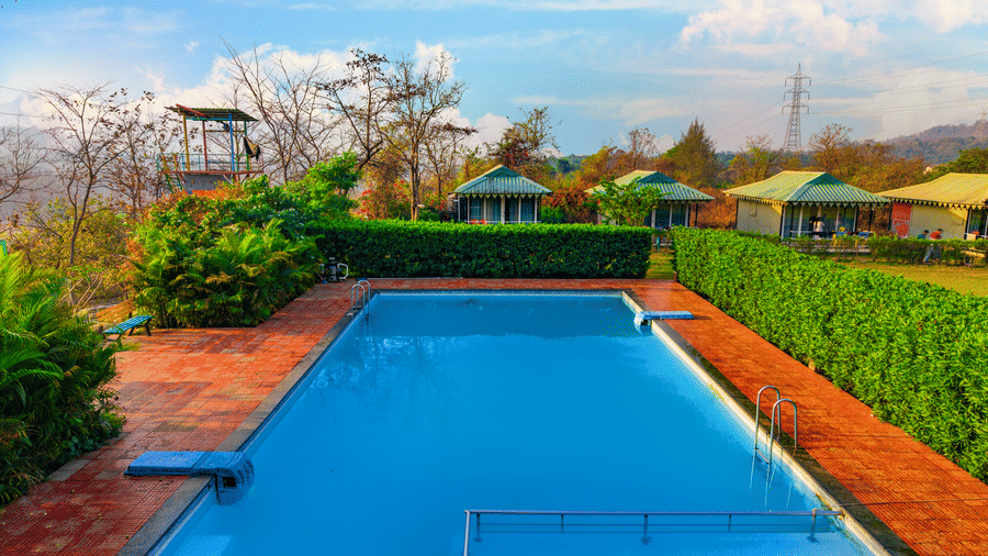 Swimming pool at Nature Trails Durshet surrounded by lush greenery, cottages, and scenic hillside views under a bright blue sky.