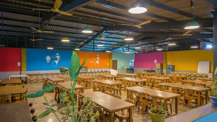 Resort dining hall interior with wooden tables and benches, colorful multicoloured accent walls, potted plants, ceiling fans and bright overhead lighting.