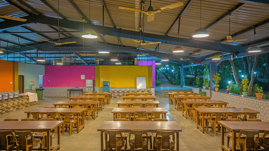 Spacious resort dining hall with rows of wooden tables and benches, vibrant yellow, pink and blue painted walls, ceiling fans and bright overhead lighting.