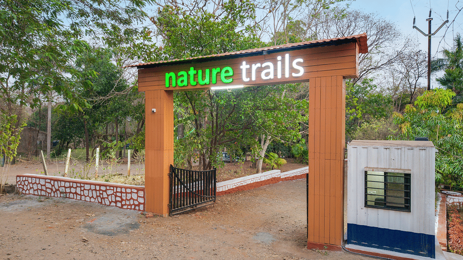 Daytime view of Nature Trails resort entrance gate with green branded signage, wooden arch frame, surrounded by trees and blue building in background.