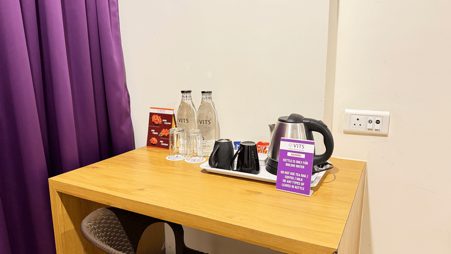 A hotel room desk with a kettle, two water bottles, and a television mounted on the wall above it at  VITS Daman Devka Beach