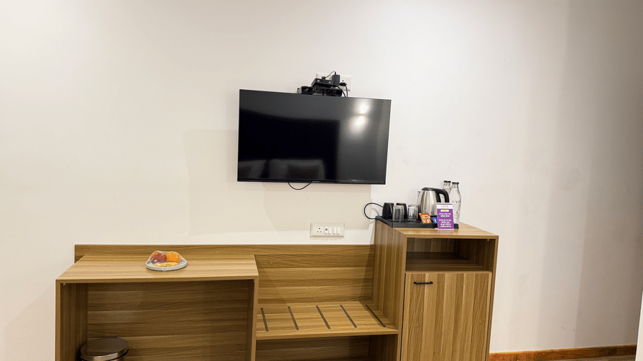 Executive Cottage at VITS Daman Devka Beach with wall-mounted TV, wooden shelf unit, kettle station, and minimal decor in a clean white-walled room.