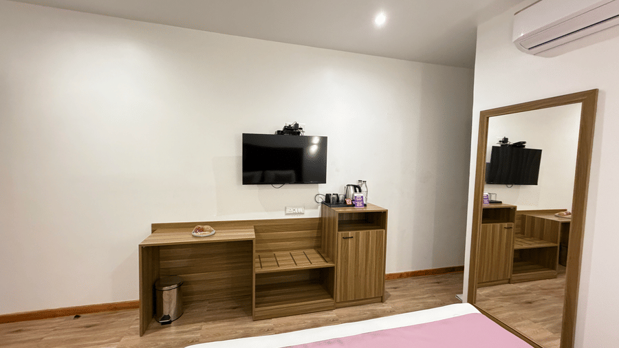 VITS Daman Devka Beach Executive Cottage workspace area with wall-mounted TV, wooden unit, full-length mirror, and kettle station against white walls.