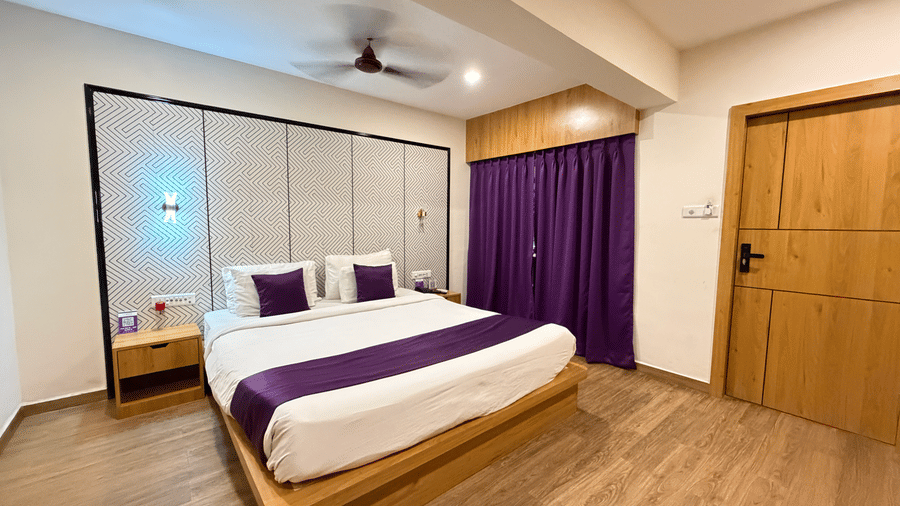  hotel room at VITS Daman Devka Beach featuring a large bed with a white and purple cover, a wooden nightstand, curtains, and light wooden flooring