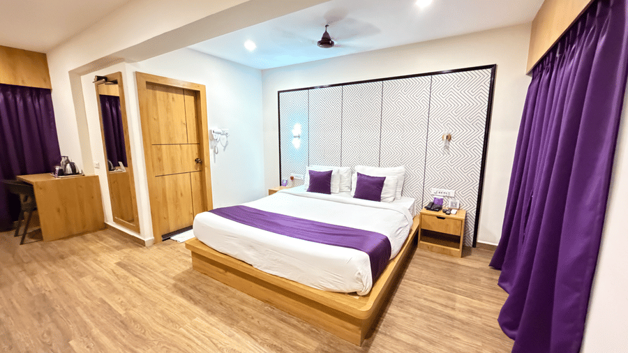 Hotel room at VITS Daman Devka Beach featuring a large bed with a white and purple cover, a wooden nightstand, curtains, and light wooden flooring.