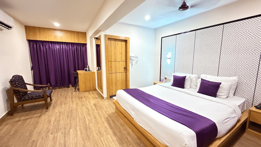 A hotel room at VITS Daman Devka Beach with a large bed dressed in white and purple linens, complemented by a wooden nightstand, soft curtains, and light wooden flooring.