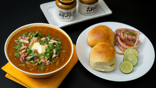 A plate filled with pav (bread), lime slices, and pickled onions, next to a big bowl with bhaji and 2 pickle jars nearby. 