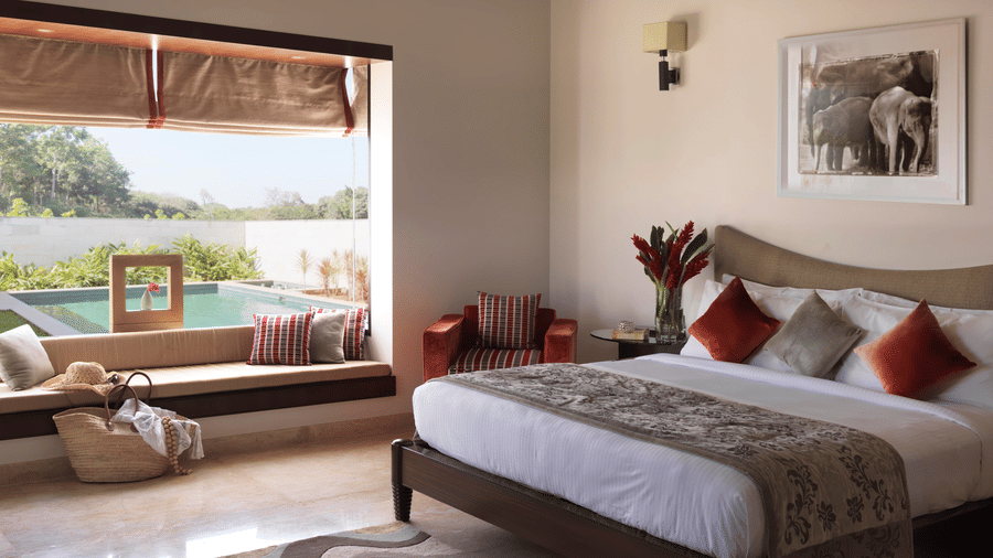 Interior of The Residence at The Serai Chikamagalur featuring a plush bed with a vase on the nightstand and next to is is a large window with seating space with a pool view